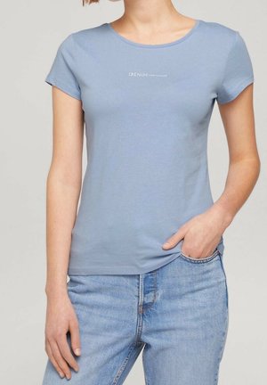 Kobieta ubrana w jasnoniebieski dopasowany t-shirt z krótkim rękawem z napisem „DENIM TOM TAILOR” i jasnoniebieskie dżinsy, jedna ręka w kieszeni.