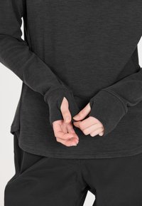 Langärmliges Oberteil in Dunkelgrau, aus weichem Material gefertigt, mit Daumenlöchern in den Bündchen und einem leicht abgerundeten Saum. Kombiniert mit schwarzen Hosen.