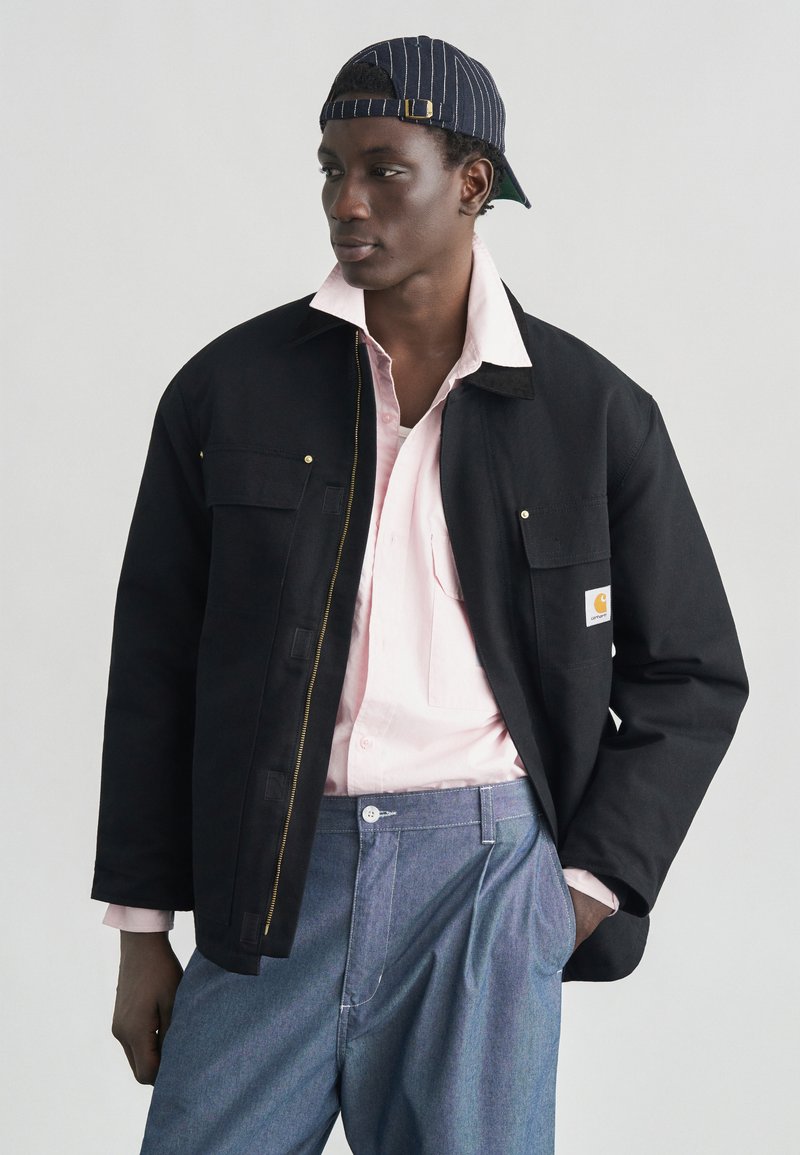 Μαύρο μπουφάν από καμβά με φερμουάρ, δύο τσέπες στο στήθος και λογότυπο Carhartt. Φορεμένο πάνω από ανοιχτό ροζ πουκάμισο και ανοιχτό μπλε παντελόνι.