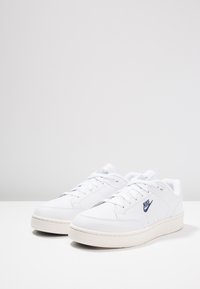 Zapatillas de piel blancas con una textura suave, con un logotipo de Nike azul, punta redondeada y una suela de goma color crema.