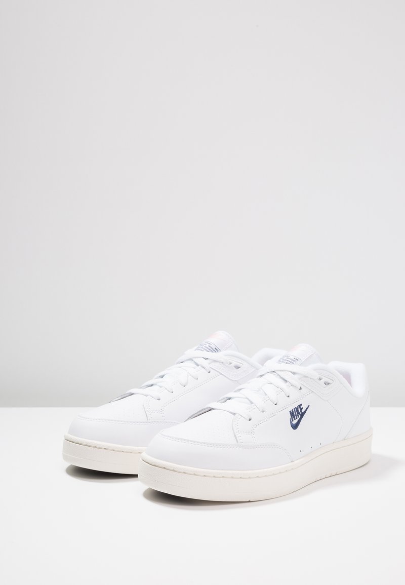 Baskets en cuir blanc avec une texture lisse, dotées d'un logo Nike bleu, d'un bout rond et d'une semelle en caoutchouc couleur crème.