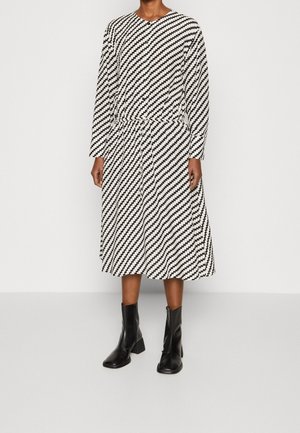 Czarna i biała diagonalna sukienka midi w zygzakowaty wzór z długimi rękawami, zestawiona z czarnymi botkami, na stojącej osobie.