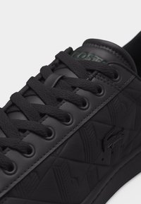 Baskets Lacoste noires avec un motif géométrique texturé, lacets noirs et logo crocodile en relief sur le côté, gros plan sur la partie avant supérieure.