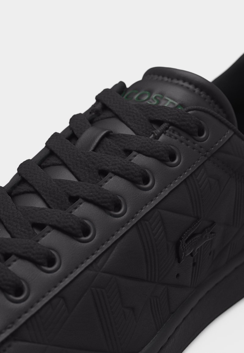 Sneaker Lacoste nera con motivo geometrico testurizzato, stringhe nere e logo del coccodrillo in rilievo sul lato, primo piano sulla parte superiore anteriore.