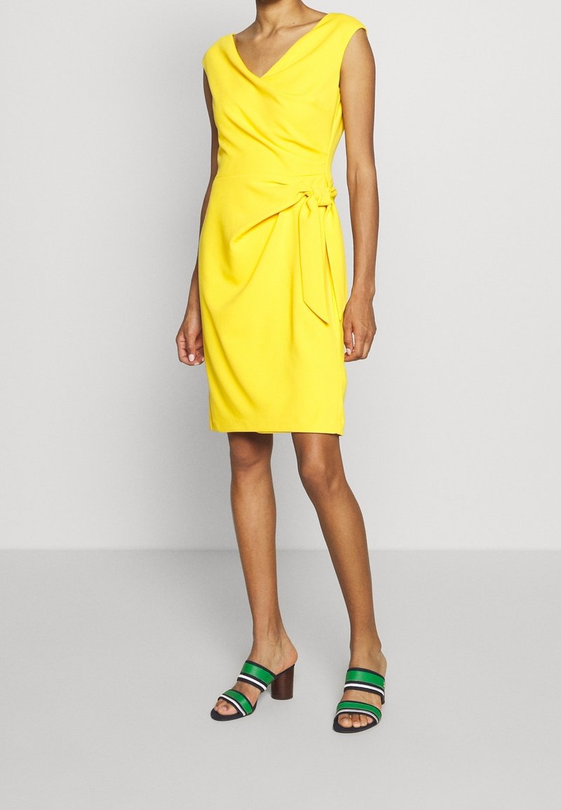 Gul ärmlös klänning med V-ringning, omslagsdesign och sidoknutdetalj. Bärs med flerfärgade randiga klackar med blockklack.