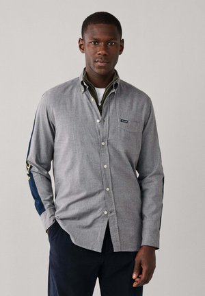 Hombre con camisa gris abotonada con rayas azul marino y letras doradas en las mangas, pantalones azul marino, de pie frente a un fondo claro y liso.
