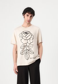 Beige t-skjorte laget av myk stoff med et stort svart blomstermotiv. Rund hals og korte ermer, med merkenavn i liten skrift.