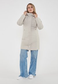 Cappotto isolato grigio chiaro con cappuccio, dotato di chiusura con zip, tasche laterali e un design elegante e aderente. Abbinato a jeans a zampa d'elefante blu chiaro.
