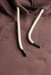 Sudadera marrón con cordones crema, que presenta un patrón de logo texturizado. El material es suave, con una textura acogedora y ligeramente pelusa.