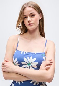 Νεαρή γυναίκα με μακριά ξανθά μαλλιά, φορώντας μπλε τοπ με λουλούδια και λεπτές τιράντες, σταυρωμένα χέρια, κοιτάζει απαλά την κάμερα μπροστά σε απλό φόντο.