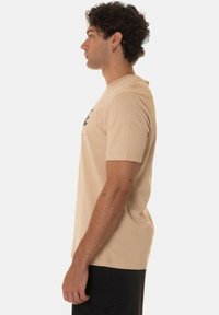 Maglietta di cotone beige a maniche corte, con una grafica sulla parte anteriore, indossata con pantaloni cargo neri. Vista laterale.