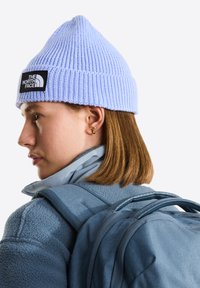 Gorro de punto azul claro con textura acanalada y parche negro con logo, llevado con una chaqueta de forro polar azul y una mochila.