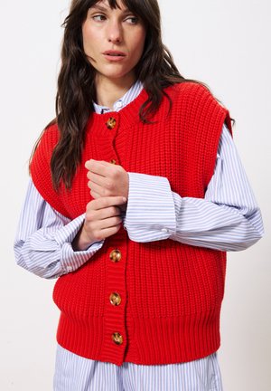 Gilet en tricot rouge avec un motif texturé, comportant de grands boutons en or. Porté par-dessus une chemise à rayures bleu et blanc avec de larges poignets.