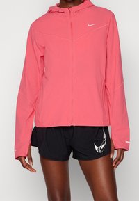 Veste légère rose avec capuche, fermeture éclair et accents réfléchissants ; associée à un short noir avec un motif graphique blanc.