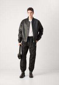 Schwarze synthetische Bomberjacke mit gesteppten Details, weißes T-Shirt und strukturierte schwarze Hose. Schwarze Stiefel und ein schwarzes Headset.