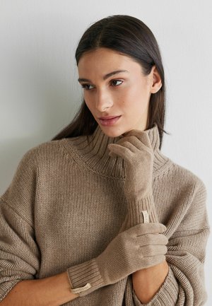 Beiga stickade handskar med ribbade manschetter och detalj med etikett, använda tillsammans med en matchande beige polotröja, mot en enfärgad bakgrund.