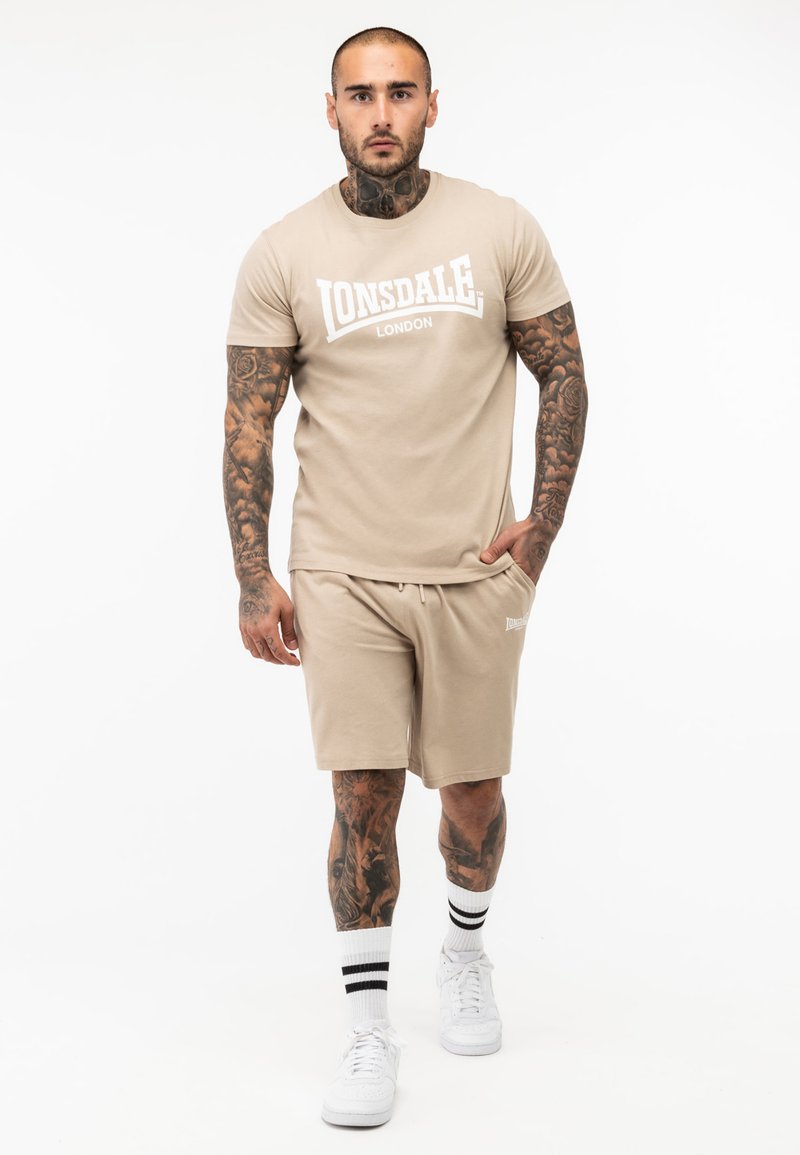 Tricou scurt bej cu imprimeu alb "Lonsdale London", asortat cu pantaloni scurți corespunzători. Afișează tatuaje, adidași albi și șosete cu dungi.