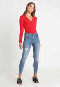 Body rosso a maniche lunghe con scollo a V rifinito in pizzo, abbinato a jeans skinny blu strappati e sandali neri con fibbia.