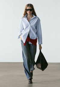 Gestreiftes blaues Hemd mit Knöpfen, lockere Jeans, roter Pullover um die Taille gebunden, schwarze Sonnenbrille und eine große schwarze Handtasche.