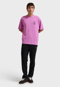 Młody mężczyzna stoi z rękami za plecami, ubrany w jaskrawy fioletowy t-shirt, czarne spodnie w wzory oraz ciemne buty sportowe na jednolitym szarym tle.