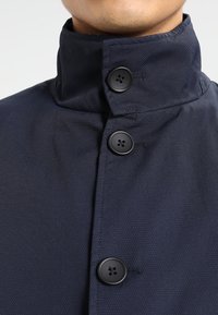 Giacca blu navy con colletto alto, tessuto cremoso e bottoni neri. Tre bottoni visibili nella zona del collo.