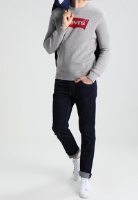 Sweat-shirt gris avec logo Levi's rouge, jeans foncés et baskets blanches. Modèle debout, tenant une veste bleu marine. Design simple, look décontracté.