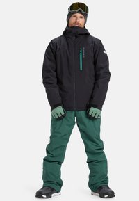 Schwarze wasserdichte Jacke mit einer Reißverschlusstasche, kombiniert mit grüner, isolierter Hose und Handschuhen. Das Modell trägt Skibrille und Stiefel.