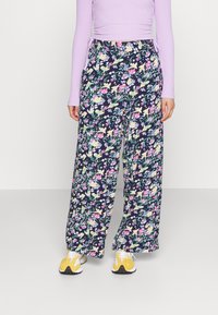 Kvinna som bär marinblå vida byxor med färgglad blommönster, lavendelfärgad långärmad topp med rynkor, och gula-vita sneakers, stående mot vit bakgrund.