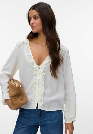 Kvinne med langt brunt hår iført en hvit bluse med volanger og blå jeans, som holder en brettet brun ruskinnveske.