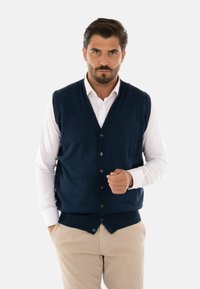 Gilet lavorato a maglia di colore blu navy con scollo a V, caratterizzato da bottoni frontali e una finitura testurizzata, indossato sopra una camicia bianca con colletto e pantaloni beige.