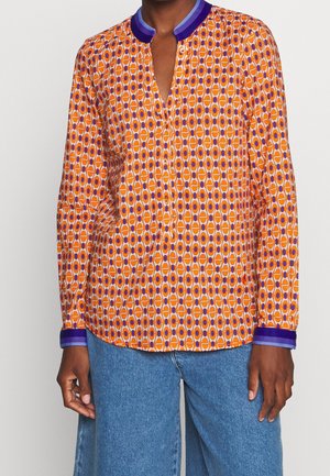 Oranje blouse met lange mouwen en patroon, met paarse kraag en manchetten, gedragen met blauwe spijkerbroek door een staand persoon.