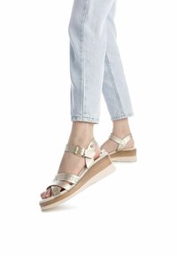 Sandalias de cuña beige con diseño de tiras recortadas, hebilla ajustable en el tobillo y un tacón de madera, combinadas con jeans de mezclilla azul claro.