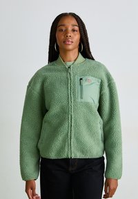 Green fleece jacket with a zip closure, textured surface, and a chest pocket in a lighter green shade. Features elasticated cuffs and collar.
