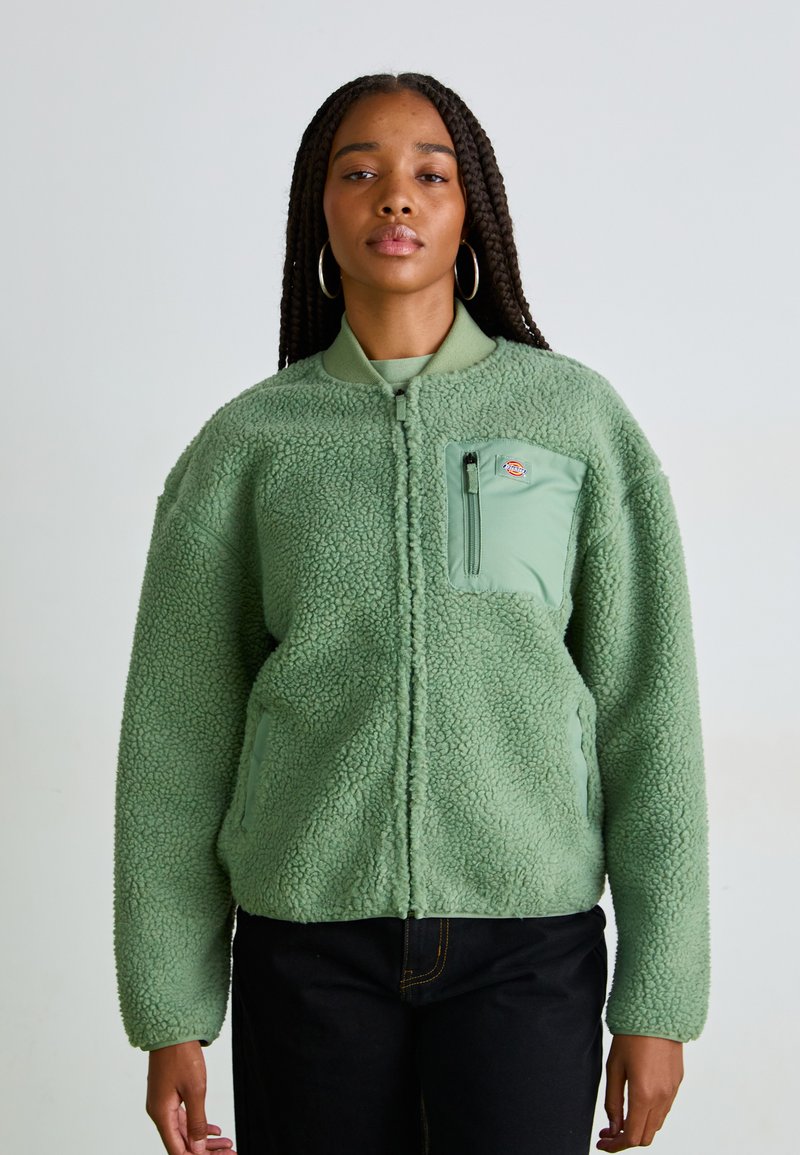 Green fleece jacket with a zip closure, textured surface, and a chest pocket in a lighter green shade. Features elasticated cuffs and collar.