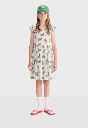 Girl wearing a green cap, white dress with orange cherry patterns, white socks, and red flip-flops, standing against a plain background.