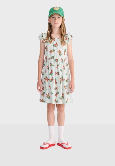 Fille portant une casquette verte, une robe blanche avec des motifs de cerises orange, des chaussettes blanches et des tongs rouges, debout devant un fond uni.