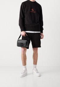 Černá mikina s červeně vyšitým logem, doplněná o černé šortky. Pod ní bílý tričko, bílé tenisky a malá černá kabelka přes rameno.