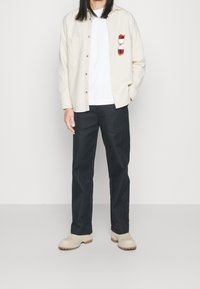 Beige paita, jossa rintatasku, mustat housut ja beiget puukengät. Valkoinen t-paita alla; aurinkolasit on työnnetty paidan taskuun.