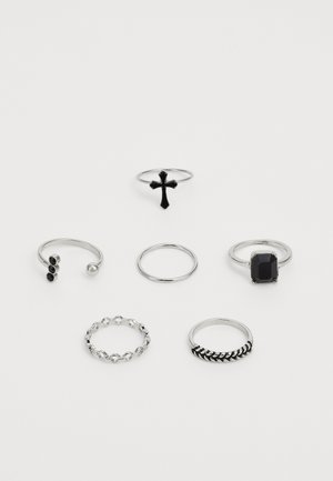 Conjunto de seis anillos en metal de tono plateado, que presentan una cruz negra, acentos texturizados y diseños variados: eslabones de cadena, piedras preciosas y bandas minimalistas.