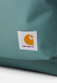 Türkiz szövet táska, amelyen egy hangsúlyos fehér folt található narancssárga logóval és a "carhartt" felirattal. A felület sima és enyhén texturált.
