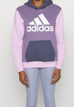 Henkilö pukeutuneena violettiin ja vaaleanpunaiseen Adidas-huppariin, jossa kengurun tasku, yhdistettynä vaaleanharmaisiin leggingsseihin, yksinkertaisen vaalean taustan edessä.