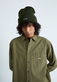 Groen geruit overhemd met een borstzak en een wit logo. Gepaard met een groene ribbelige beanie met een logopatch op de voorkant.