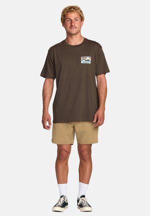 Camiseta de algodón marrón con un pequeño gráfico de montaña en azul y blanco en el lado izquierdo del pecho, combinada con pantalones cortos beige y zapatillas negras.