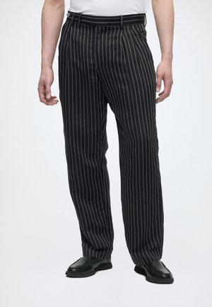 Άνδρας που φοράει μαύρο παντελόνι με ρίγες και μαύρα παπούτσια, στέκεται με τα χέρια χαλαρά στο πλάι σε λευκό φόντο.