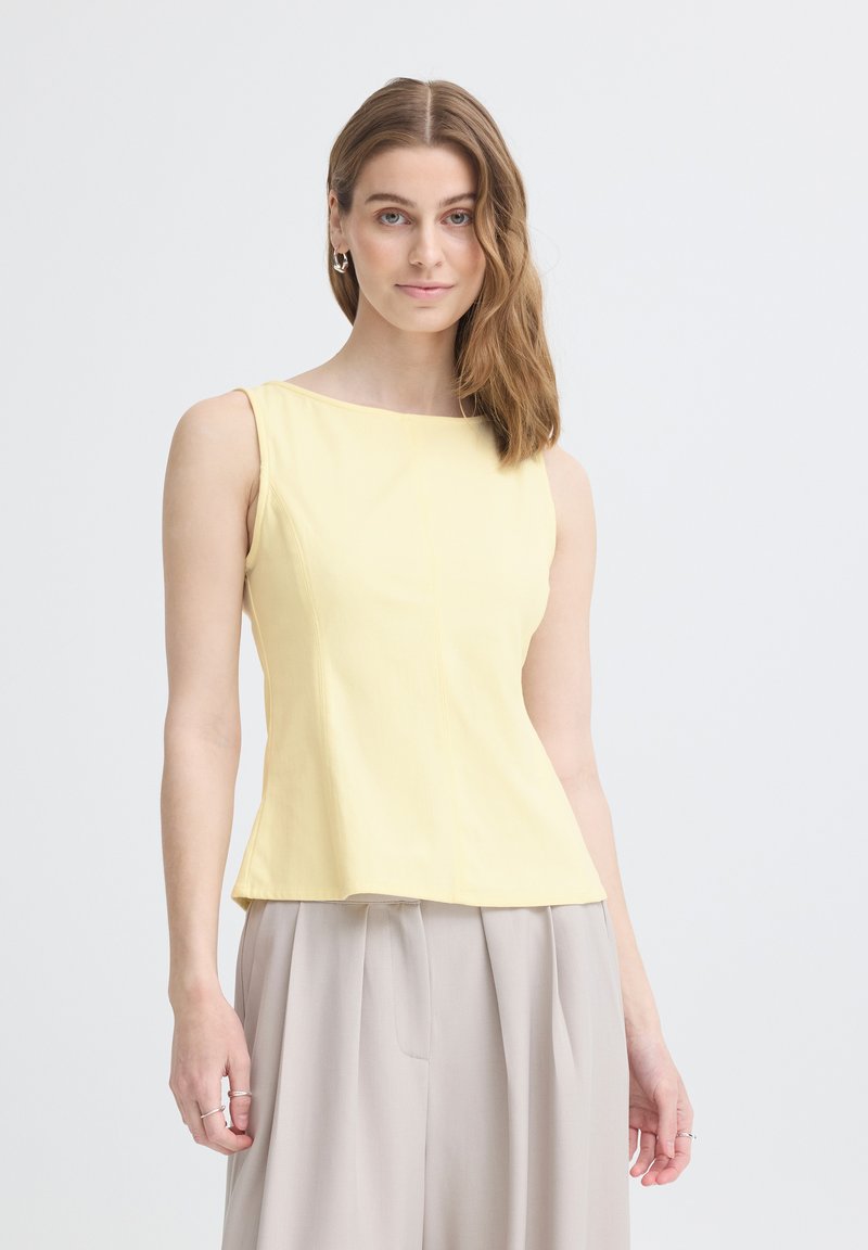Femme aux cheveux bruns mi-longs, portant un haut jaune sans manches et un pantalon plissé beige clair, debout sur un fond blanc.