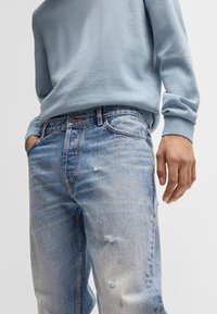 Jeans de mezclilla de lavado claro con un ajuste relajado, que presentan detalles desgastados, un desvanecimiento sutil y un diseño clásico de cinco bolsillos. Combinados con un suéter azul.