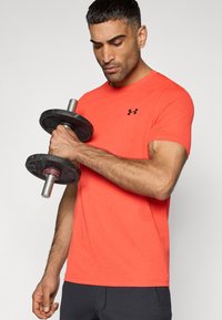 Αθλητική κοντομάνικη μπλούζα σε φωτεινό κοραλλί με υφή και μικρό μαύρο λογότυπο, συνδυασμένη με σκούρο σορτς, κρατώντας ένα αλτήρα.