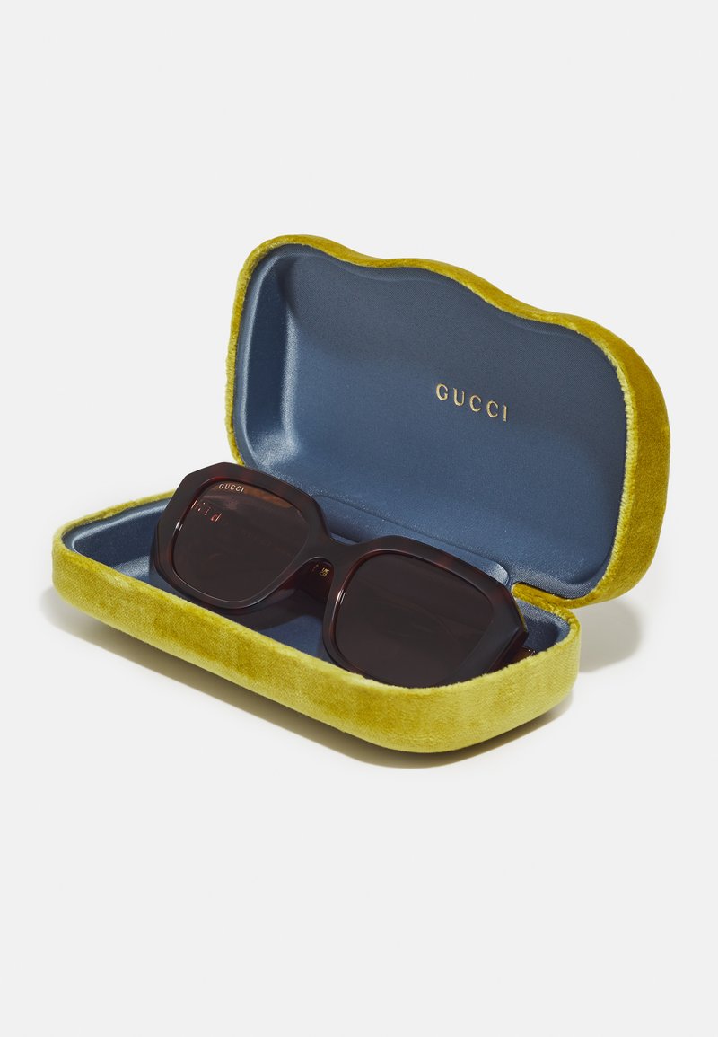 Lunettes de soleil Gucci dans une monture rectangulaire en ambre, présentées dans un étui en velours vert avec un intérieur bleu et un logo doré.