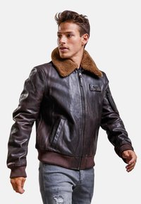 Veste bombardier en cuir marron avec un col en fourrure, des poignets côtelés, des poches zippées et un patch logo sur la poitrine. Portée sur un jean gris déchiré.