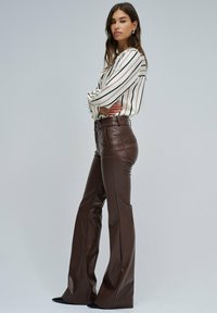 Vrouw die met gekruiste armen staat, gekleed in een gestreepte blouse en hooggepaste bruine leren broeken, tegen een effen grijze achtergrond.